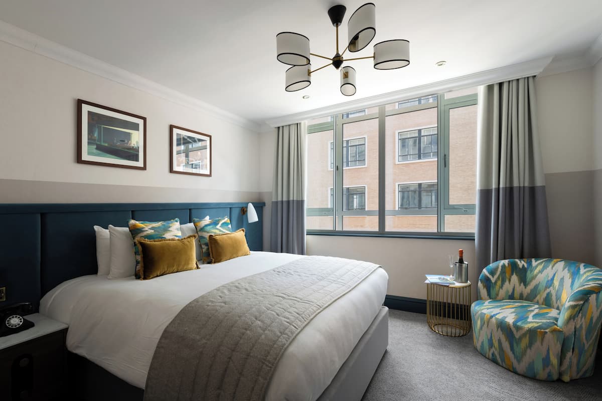 Quarto de hotel com uma cama grande, uma cadeira moderna, uma mesa lateral com abajur e vista para um edifício pela janela.