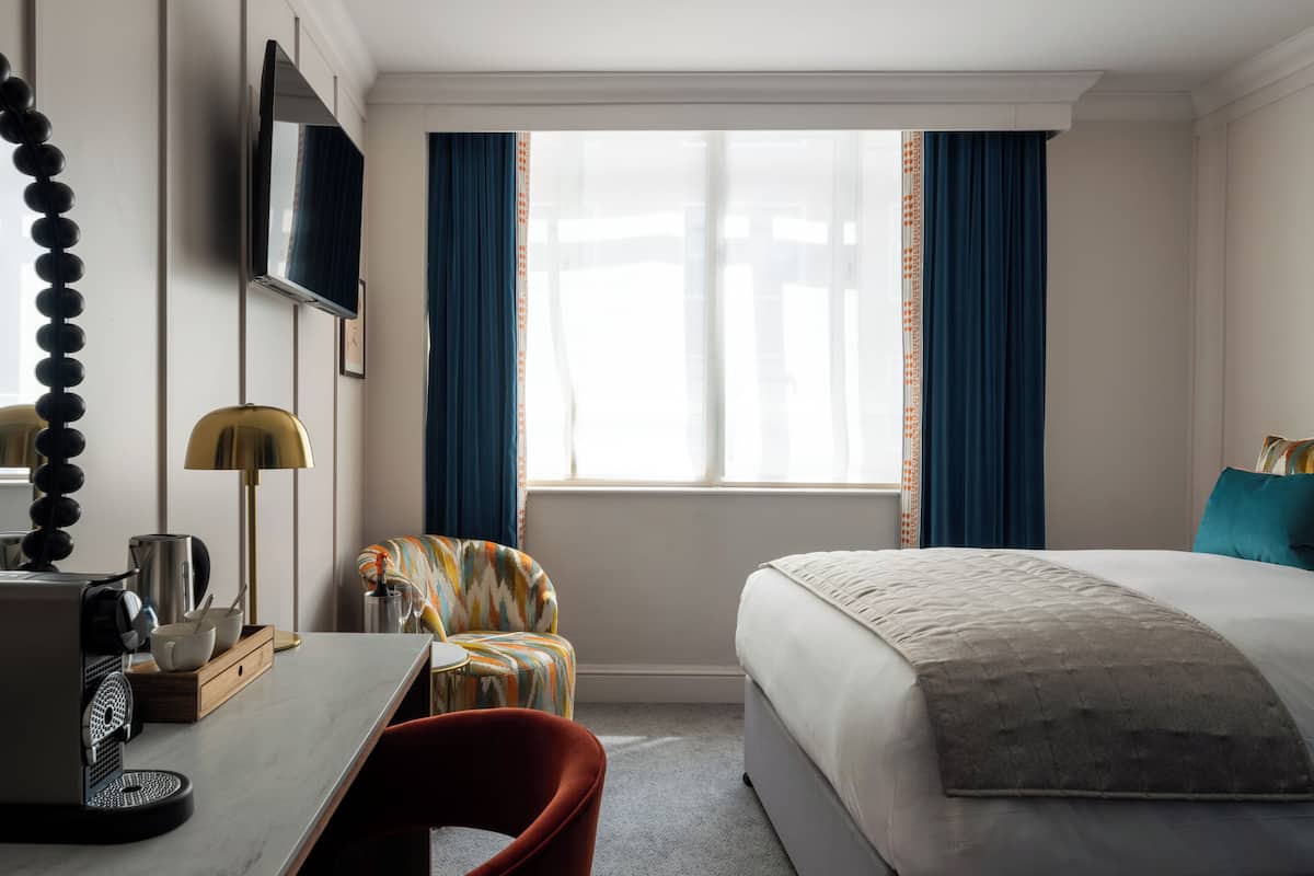 Quarto de hotel com cama, escrivaninha com cafeteira, poltrona e televisão na parede.