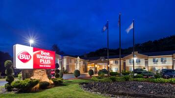 A Best Western PLUS Inn & Suites with a well-lit sign, multiple flags, and a landscaped entrance.