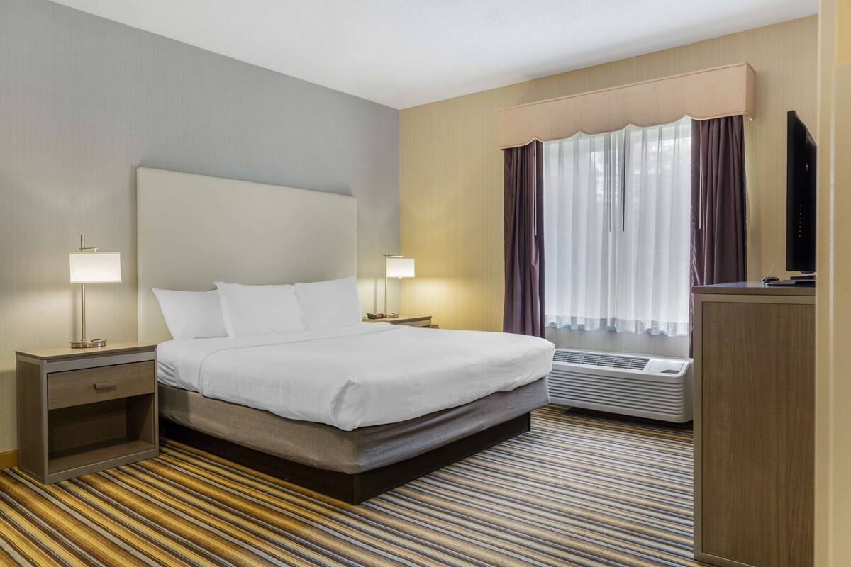 A hotel room with a large bed, two bedside tables with lamps, a flat-screen TV, an air conditioning unit, and a window with curtains.