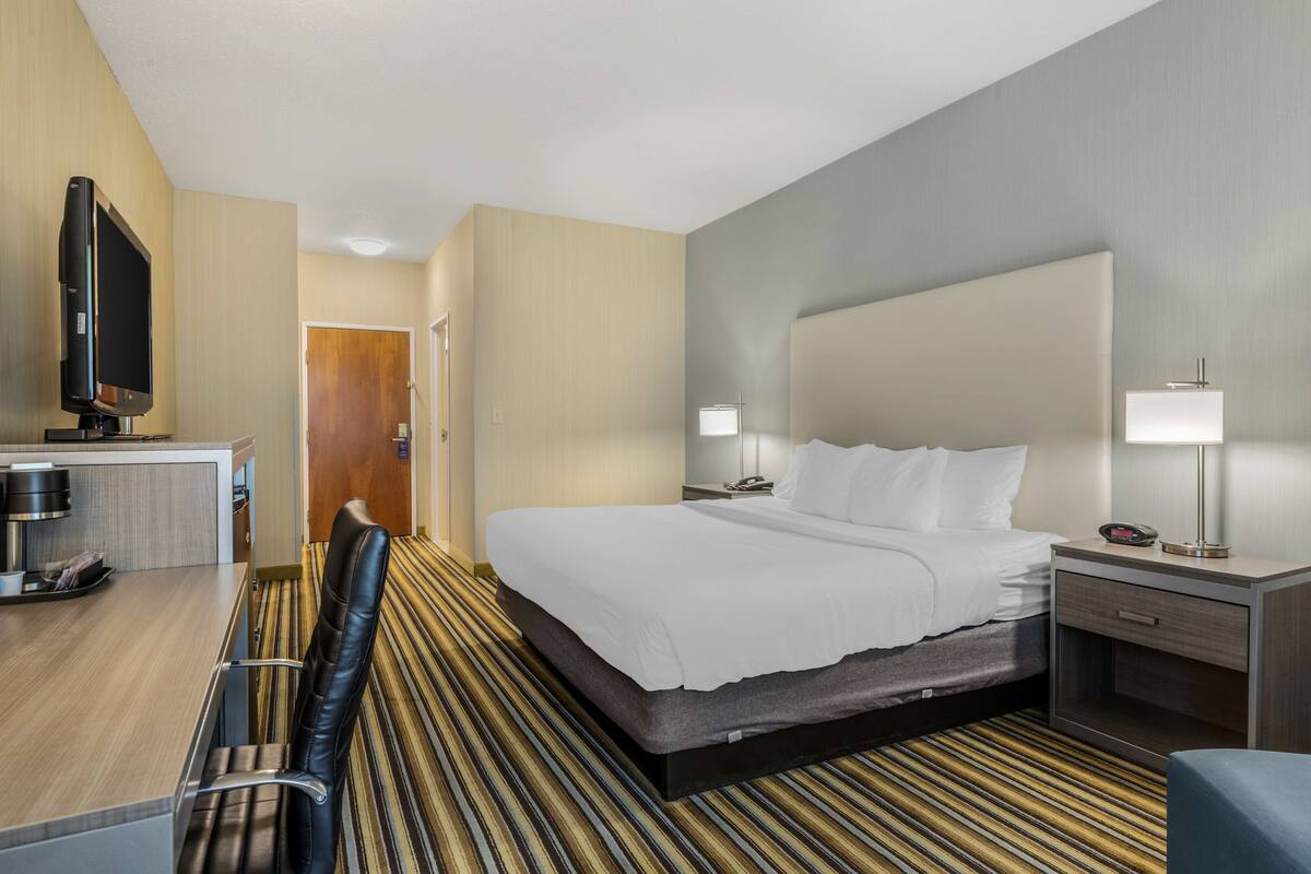 A hotel room with a bed, bedside tables, a desk with a chair, a television, and a door leading to another room.