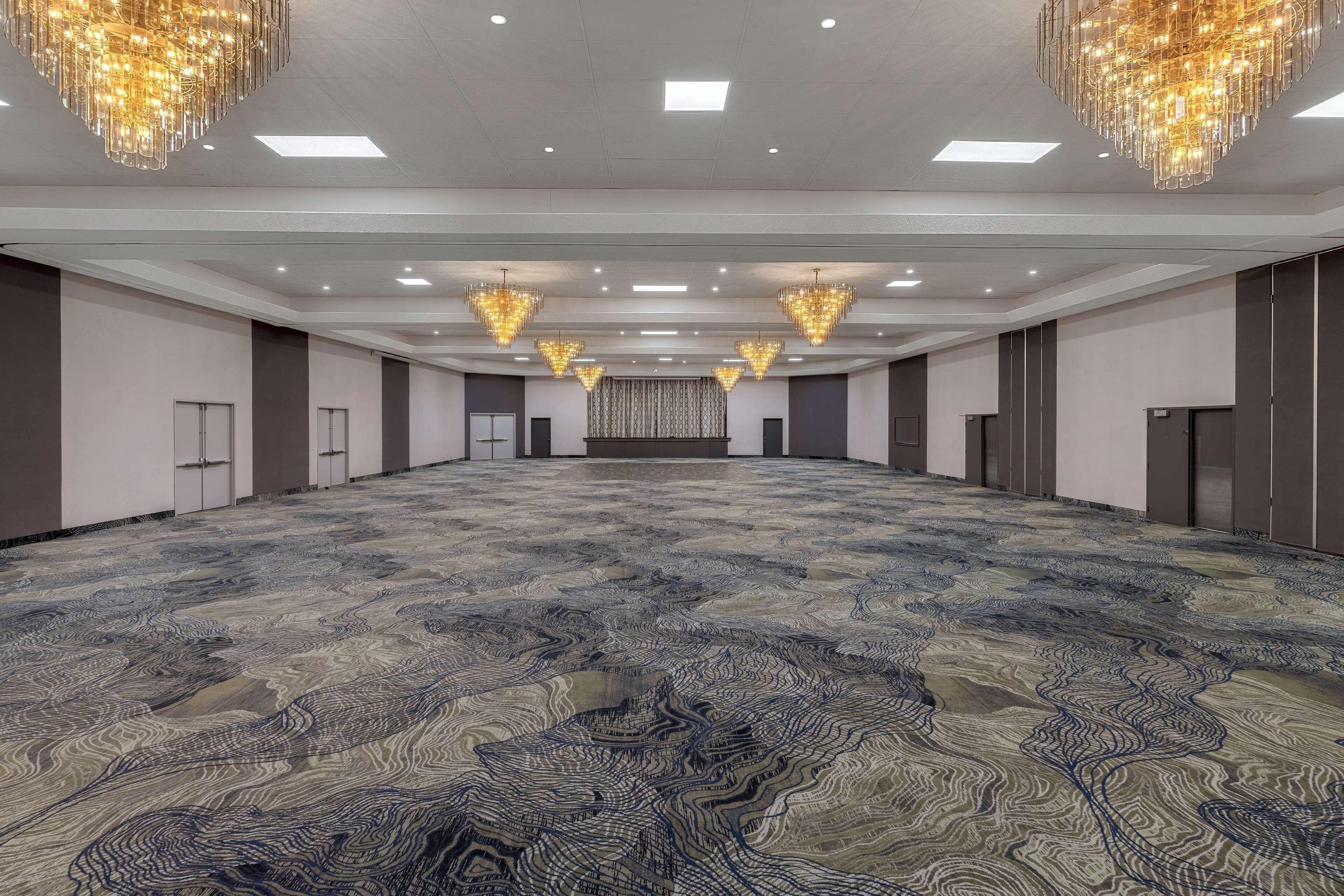 Un salón de conferencias amplio y vacío con alfombra estampada, varios candelabros y una fila de puertas a un lado.