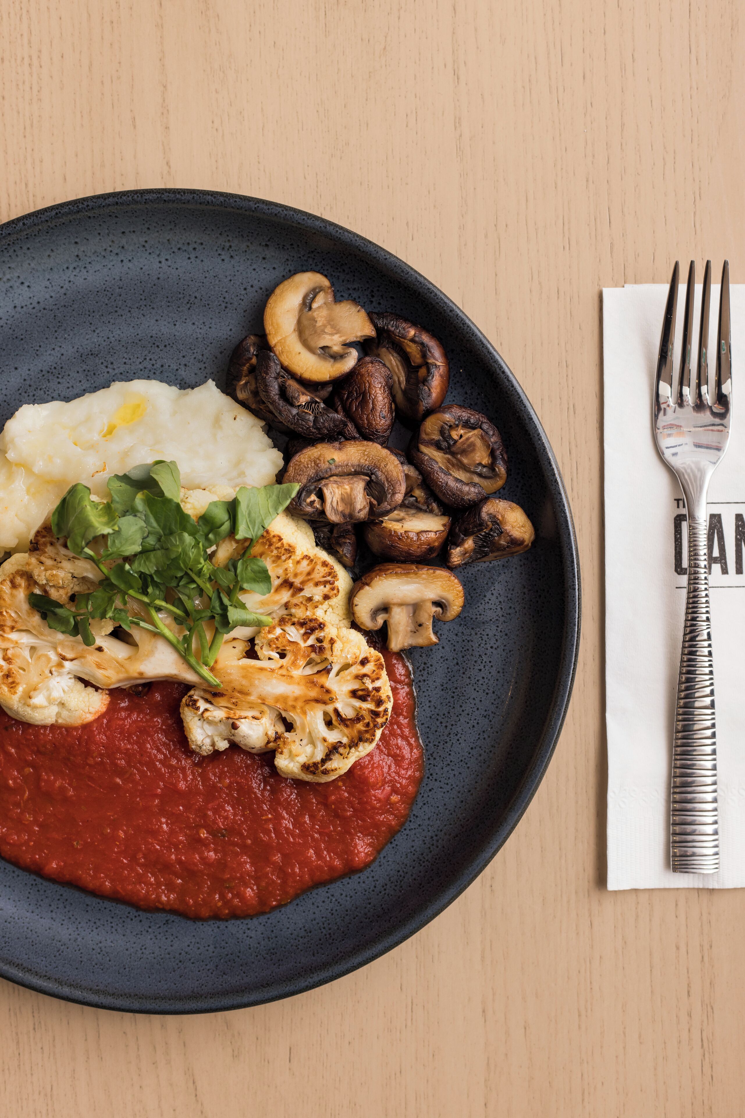 A plate with mashed potatoes, grilled mushrooms, and a red sauce, accompanied by a fork and napkin on a wooden table.