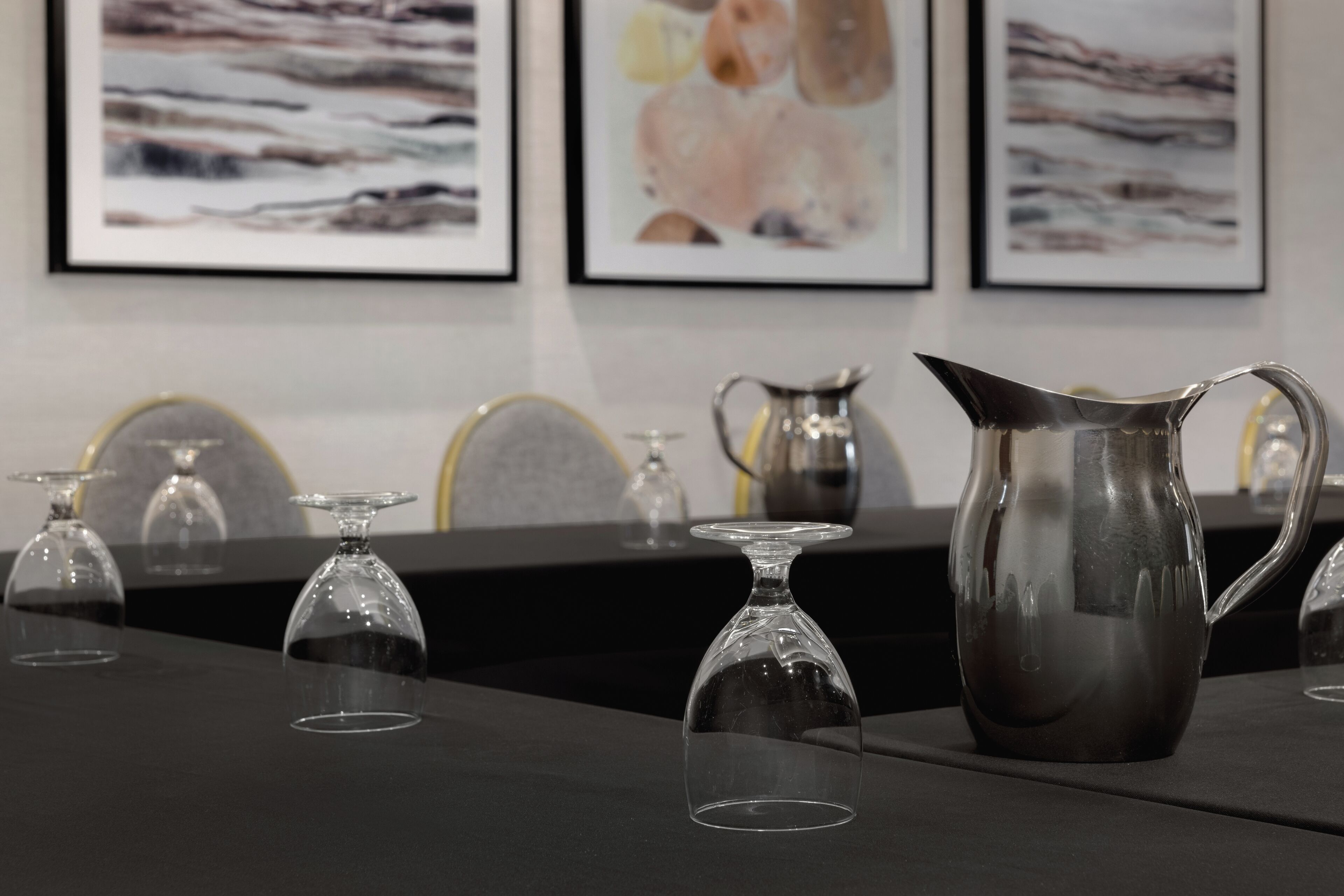 A table with a pitcher and wine glasses, framed artwork on the wall, and a black tablecloth.