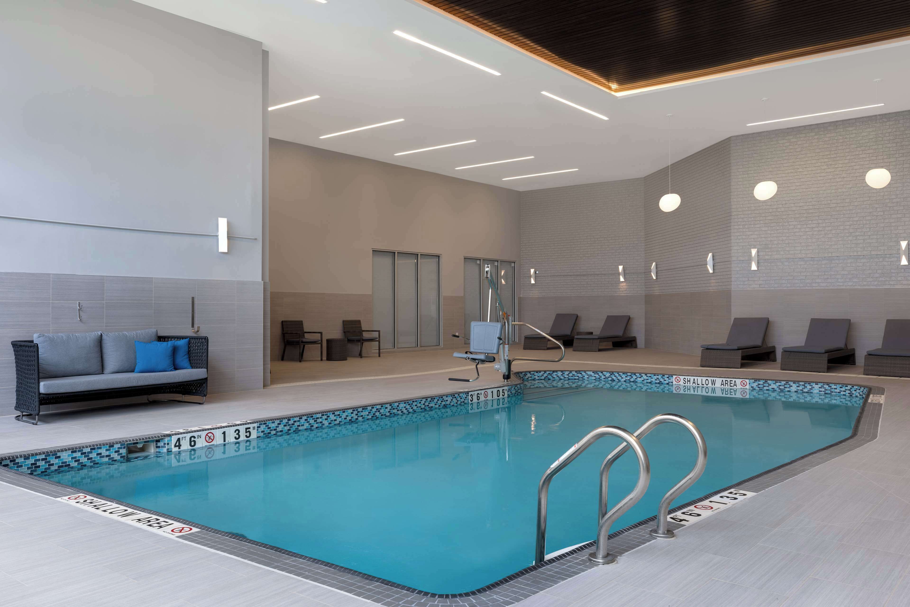 A modern indoor swimming pool with clear water, a lounge area with chairs, and a sofa.