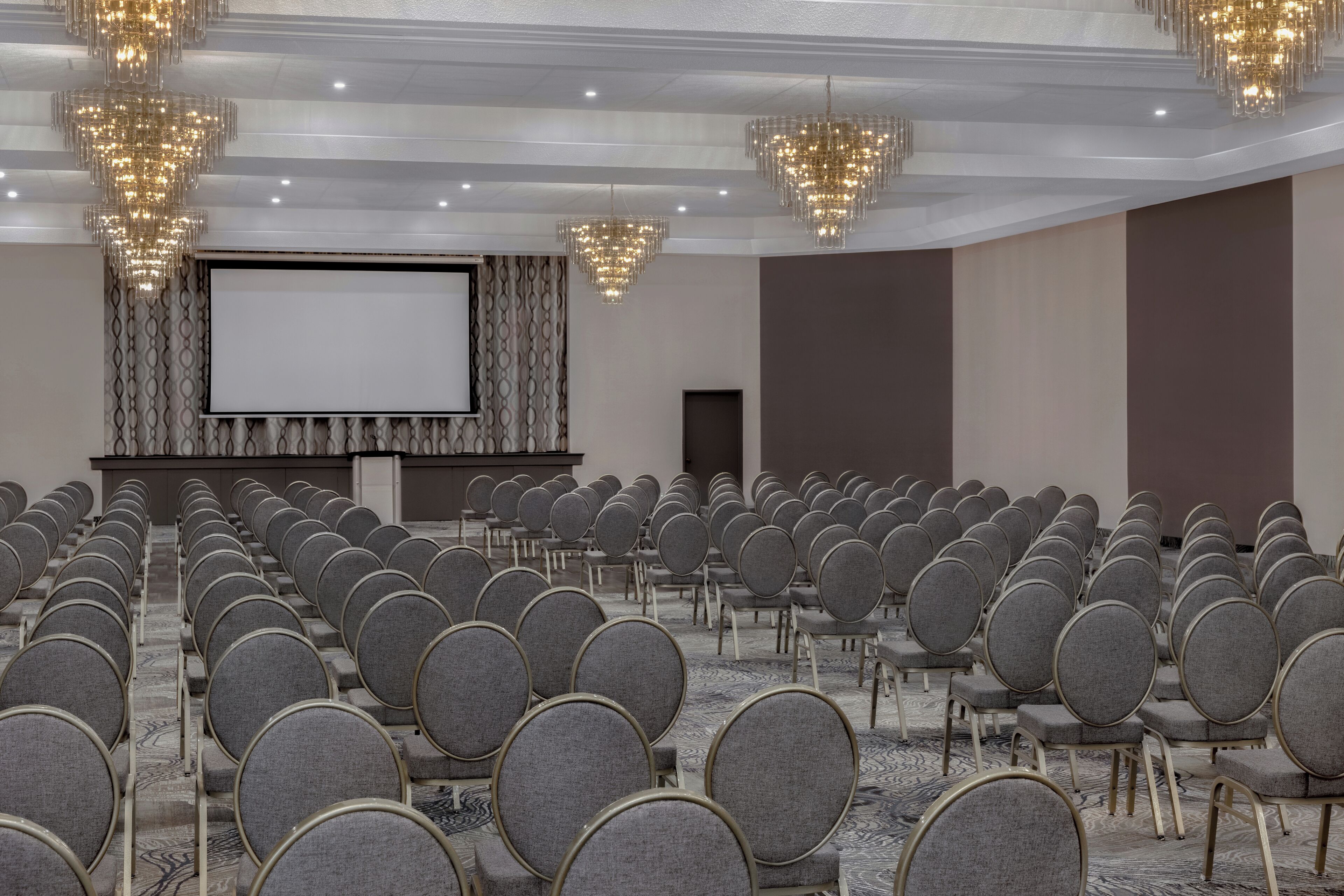 Ein Konferenzraum mit Reihen grauer Stühle, einem großen Bildschirm und Kristallleuchtern.