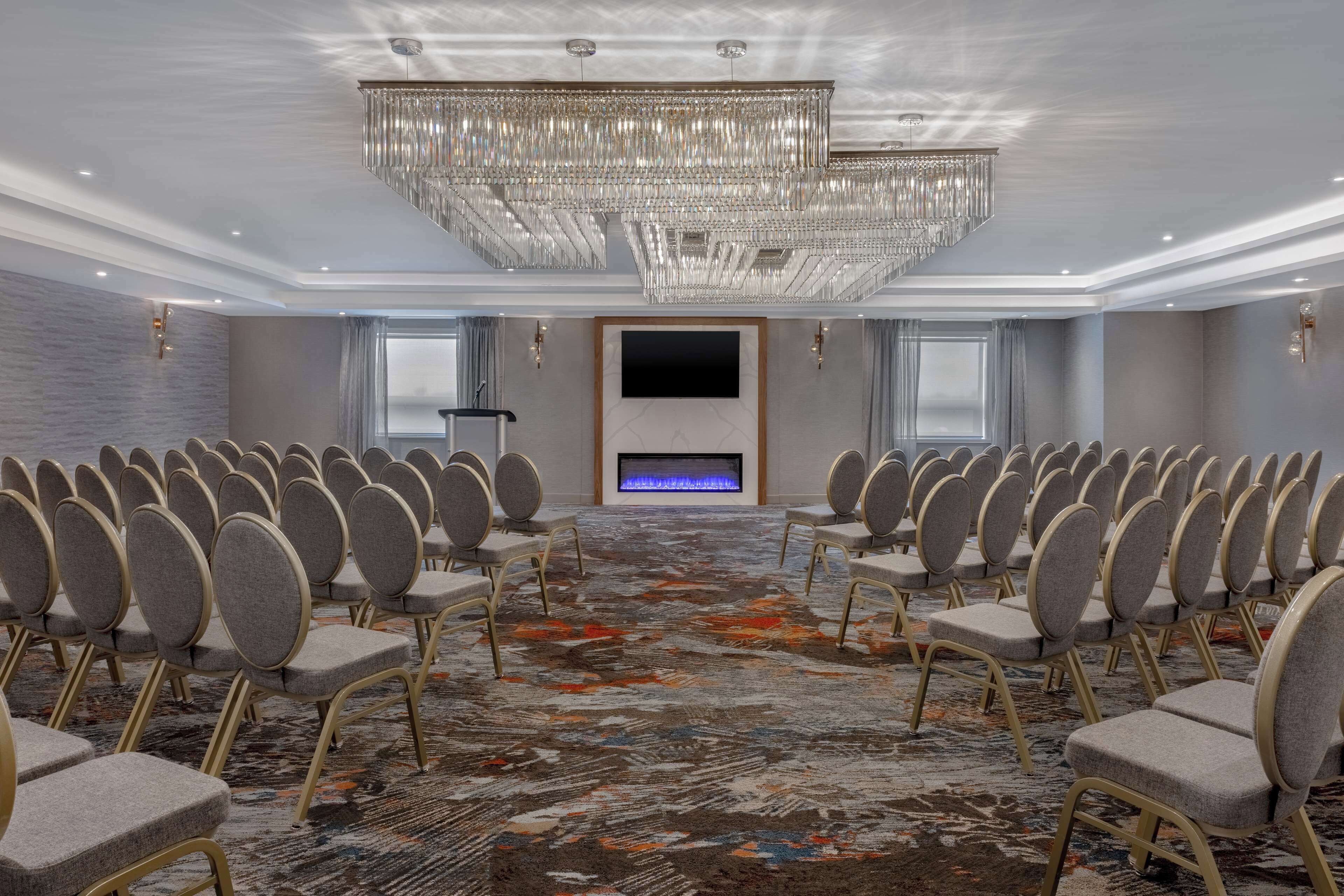A conference room with rows of chairs, a large chandelier, a flat-screen TV, and a patterned carpet.