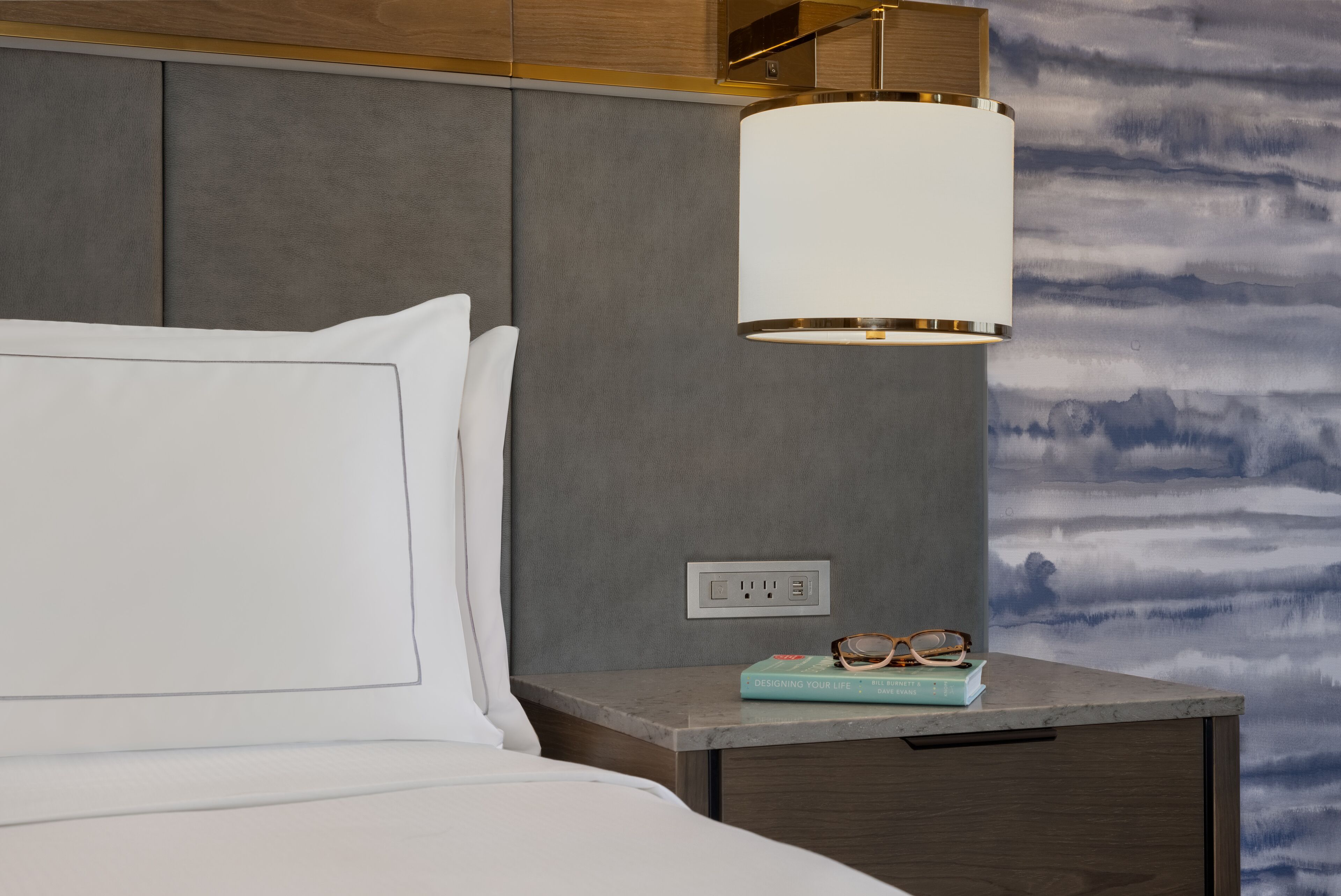 A neatly made bed with white linens, a bedside table with a lamp, and a book and glasses on it.