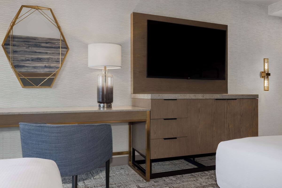 A hotel room with a wooden desk, a flat-screen TV mounted above it, a modern lamp, and a large mirror with a geometric frame.