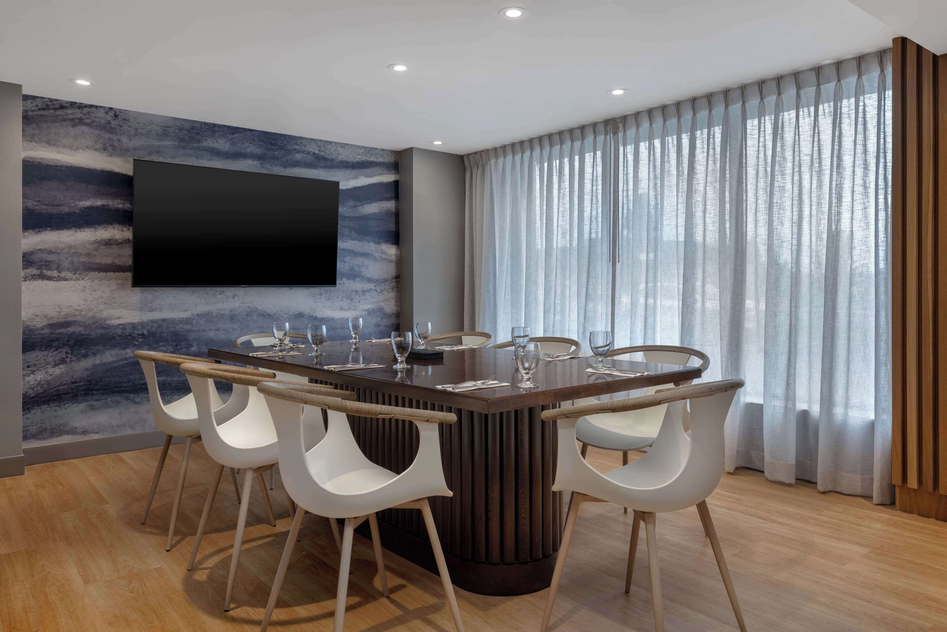 A modern dining area with a central table, white chairs, a mounted TV, and a large window with sheer curtains.