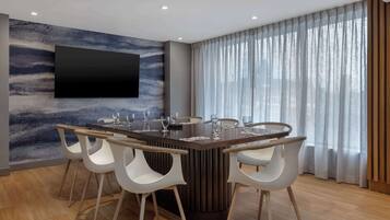 A modern dining area with a central table, white chairs, a mounted TV, and a large window with sheer curtains.