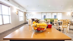 Un hall de hotel con una isla central que presenta un frutero, zona de estar con mesas y sillas, y ventanas amplias que aportan luz natural.
