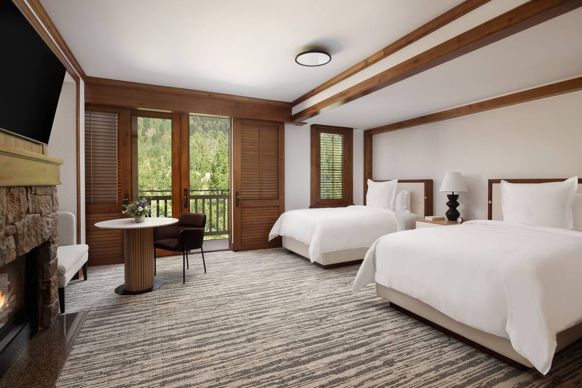 A hotel room with two beds, a fireplace, a balcony with wooden shutters, and a dining area with a table and chairs.