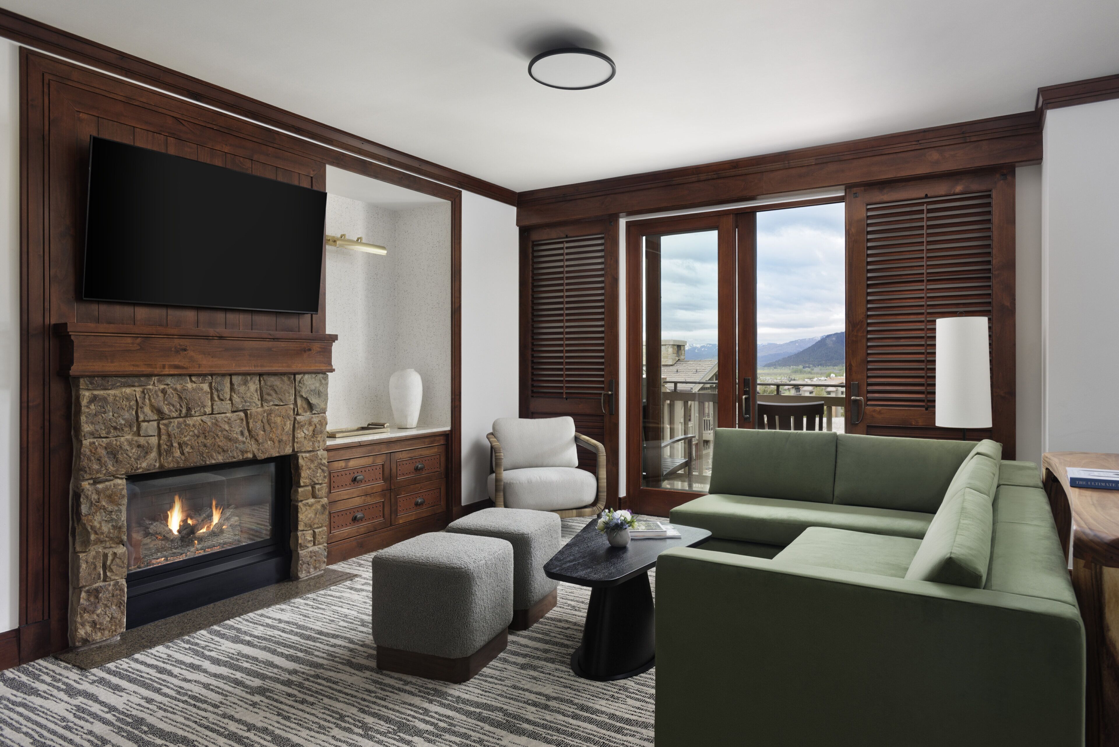 A modern living room with a stone fireplace, a flat-screen TV, a green sofa, and a view of mountains through large windows.