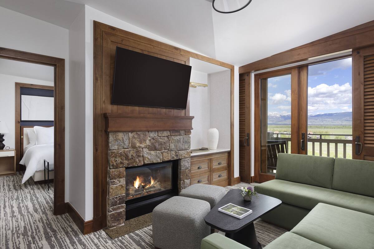 A room with a stone fireplace, a flat-screen TV mounted above it, a green sofa, and a view of the outdoors through large windows.