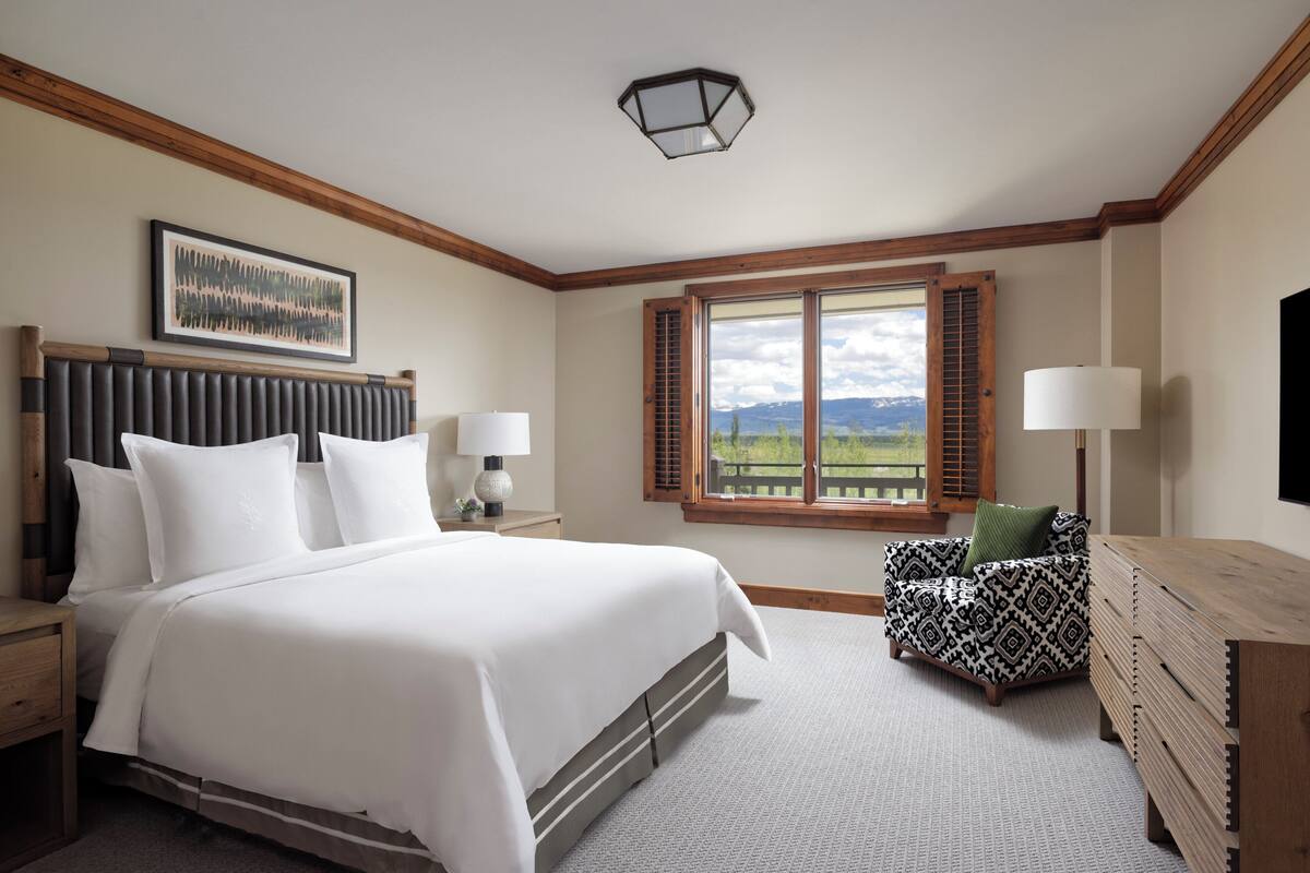 A bedroom with a large bed, a wooden headboard, a nightstand, a dresser, a window with shutters, and a view of mountains.