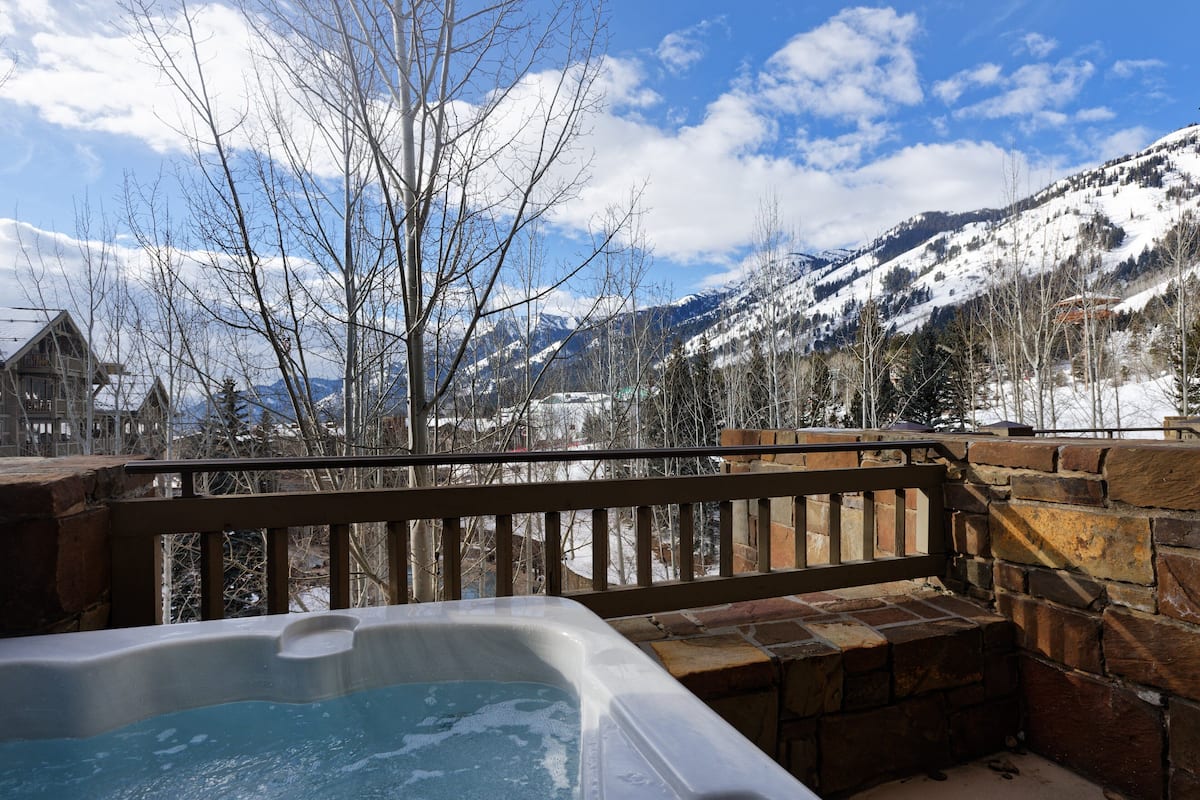 A hot tub with a stone wall and wooden railing, overlooking a snowy mountain landscape.