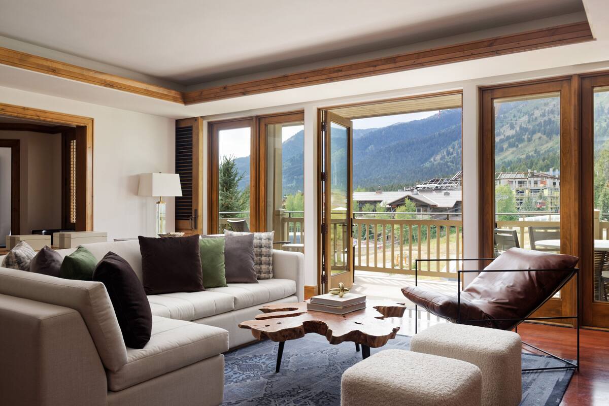 A spacious living room with a sofa, a leather chair, and a coffee table. Large windows offer a view of mountains and a balcony.