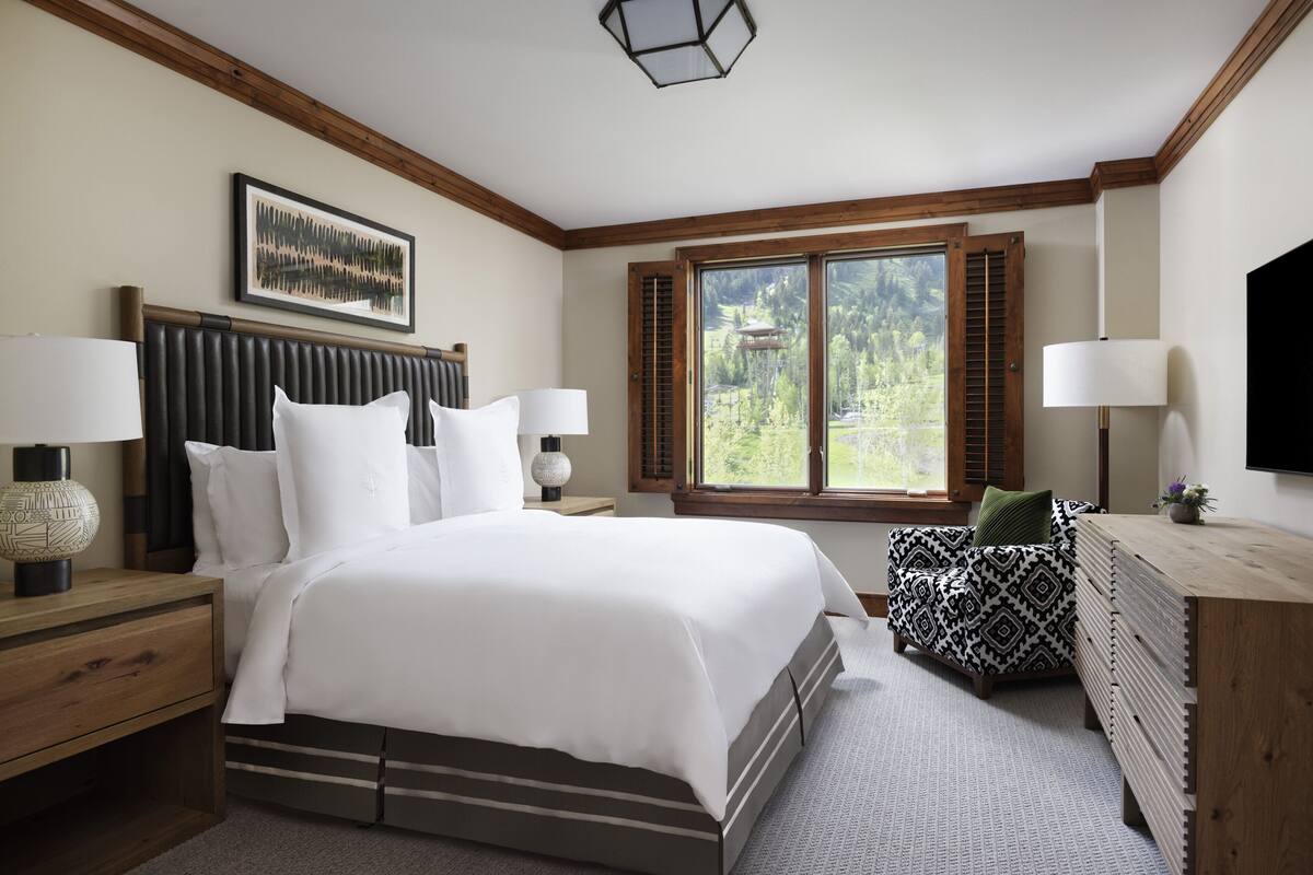 A bedroom with a bed, two bedside tables, a dresser, a window with shutters, and a ceiling light fixture.