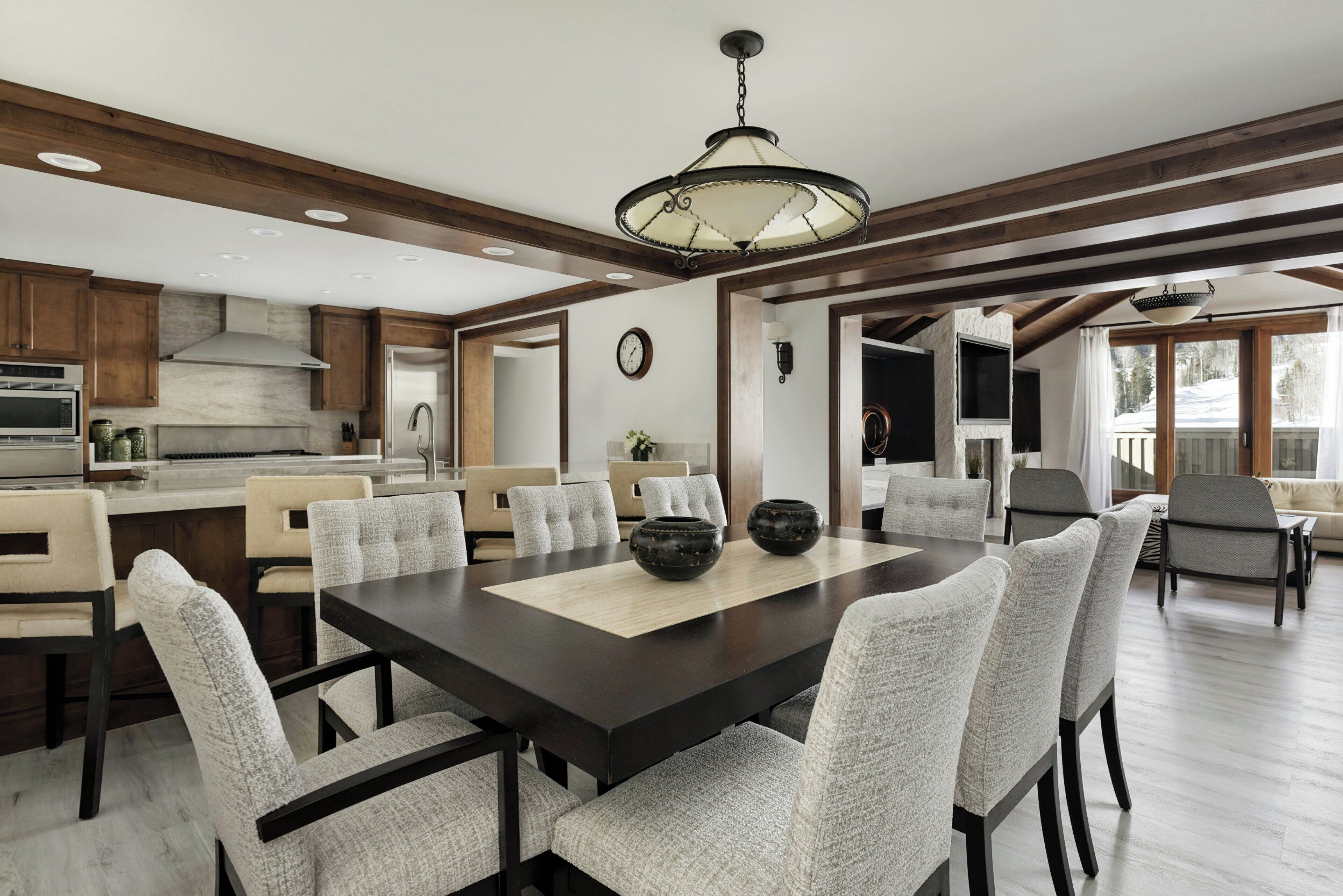A spacious dining area with a large wooden table and upholstered chairs, a kitchen with modern appliances, and a view of the outdoors through large windows.