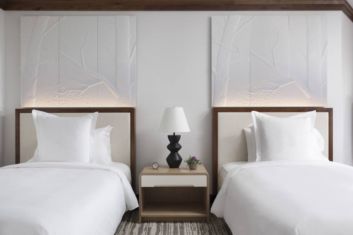 A hotel room with two beds, a nightstand with a lamp, and a wall with a tree design.