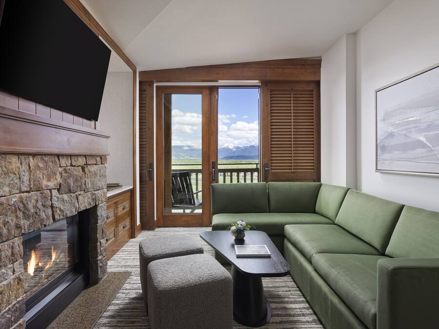Uma sala de estar moderna com lareira de pedra, um sofá verde, uma mesa de centro e uma grande janela com vista para as montanhas.