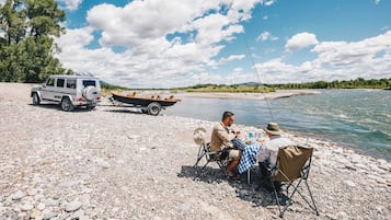 A white SUV parked on a rocky shore with a small boat nearby. Two people are seated at a table with a hat on the ground.