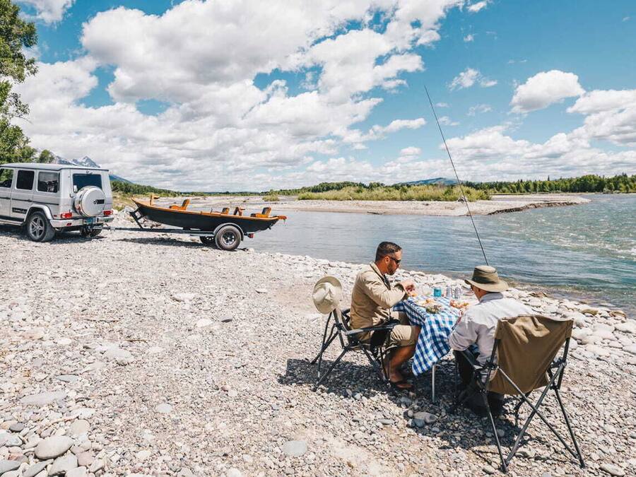 A white SUV parked on a rocky shore with a small boat nearby. Two people are seated at a table with a hat on the ground.