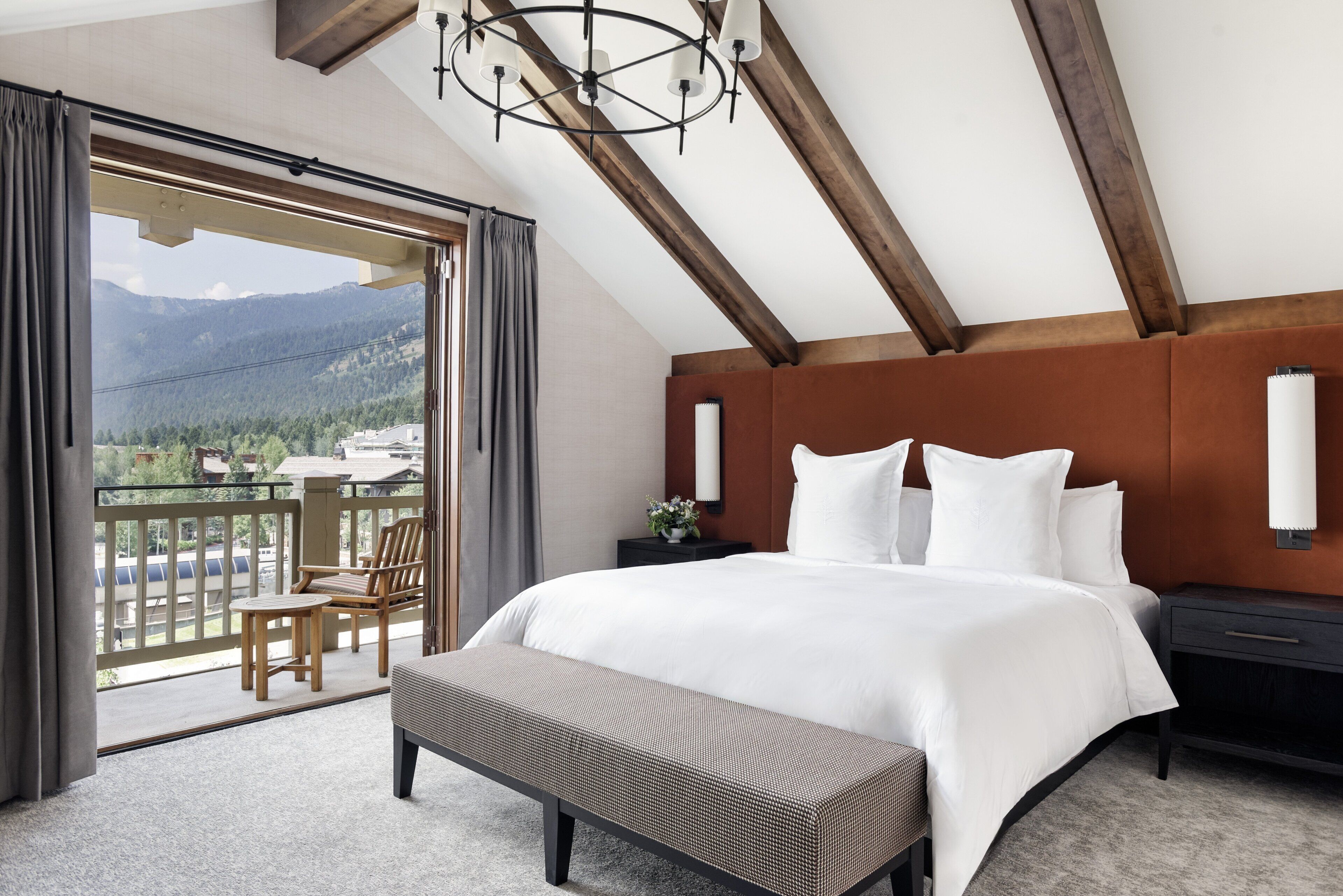 A bedroom with a large bed, a bench, a nightstand, and a view of mountains through a balcony.
