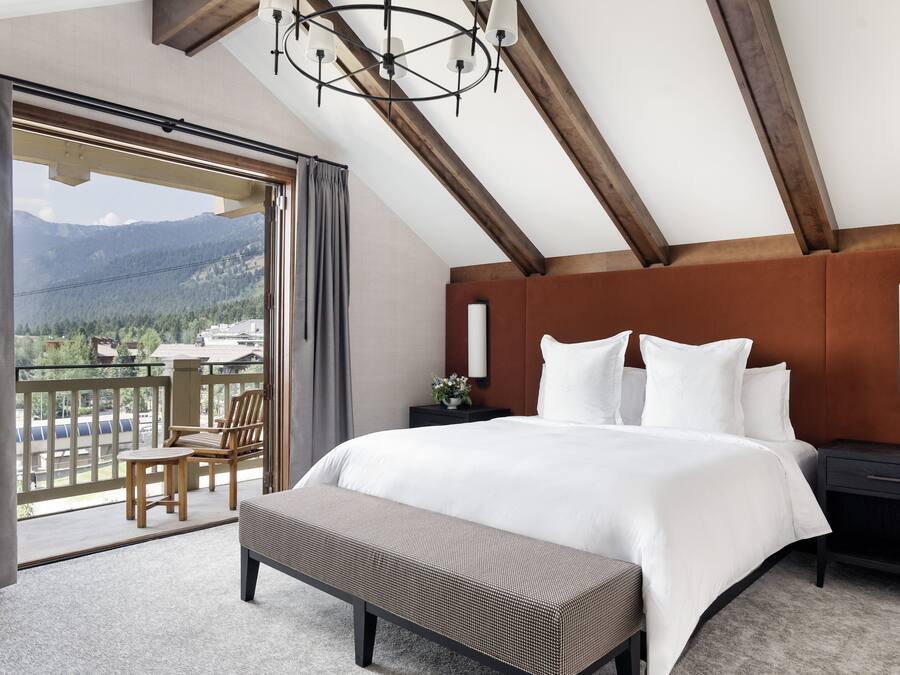 A bedroom with a large bed, a bench, a nightstand, and a view of mountains through a balcony.