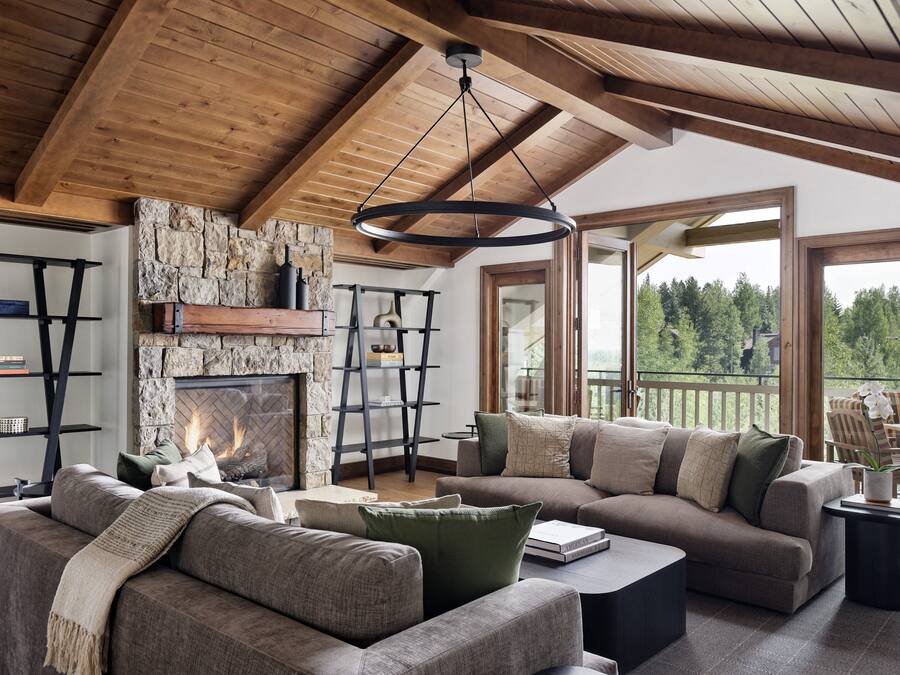 A cozy living room with a stone fireplace, wooden beams, and large windows offering a view of the outdoors.
