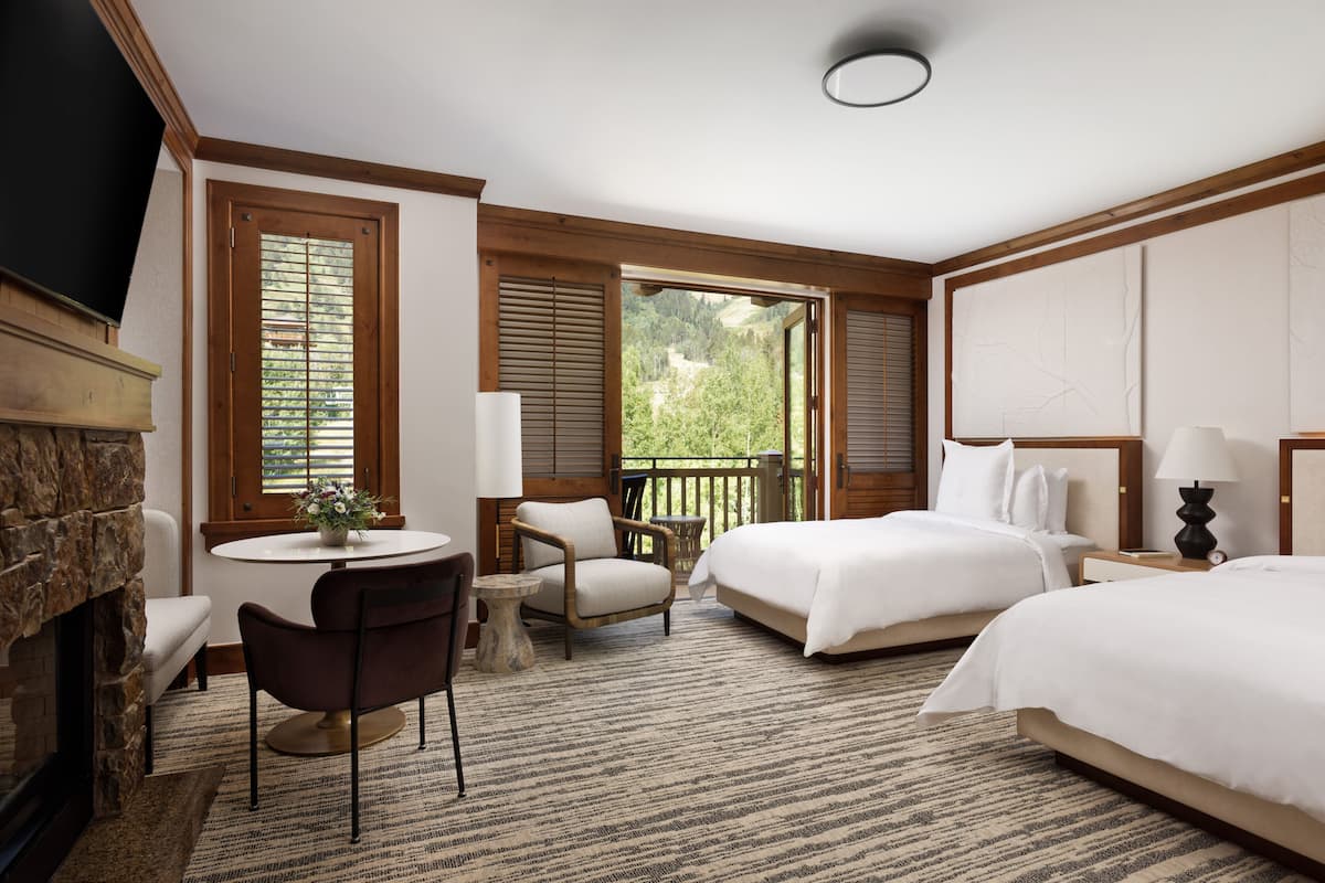 A hotel room with a fireplace, two beds, a dining table with chairs, and a balcony with a view of greenery.