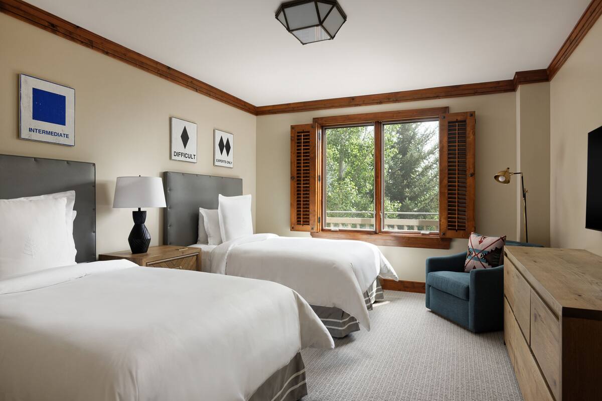 A hotel room with two beds, a wooden headboard, a desk with a lamp, a chair, and a large window with shutters.