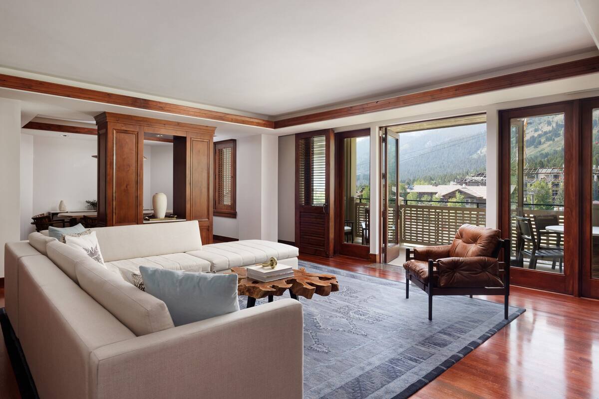 A spacious living room with a large sofa, a coffee table, and a leather armchair. The room has wooden flooring, large windows, and a view of a mountainous landscape.