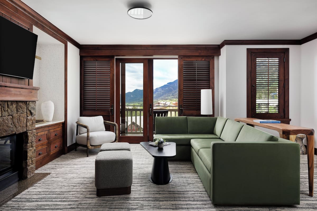 A living room with a stone fireplace, a green sofa, a wooden coffee table, and a view of mountains through large windows.