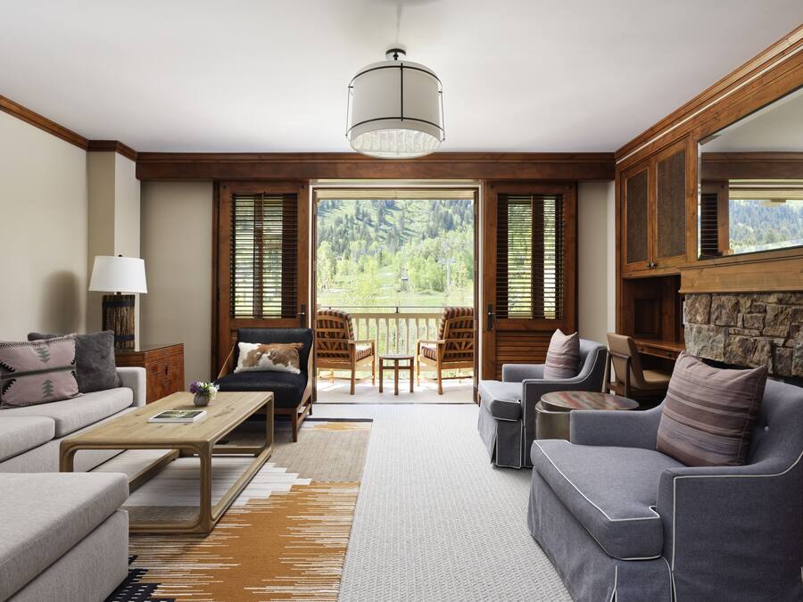 A spacious living room with a sofa, armchairs, and a coffee table. A large window offers a view of a forested area.