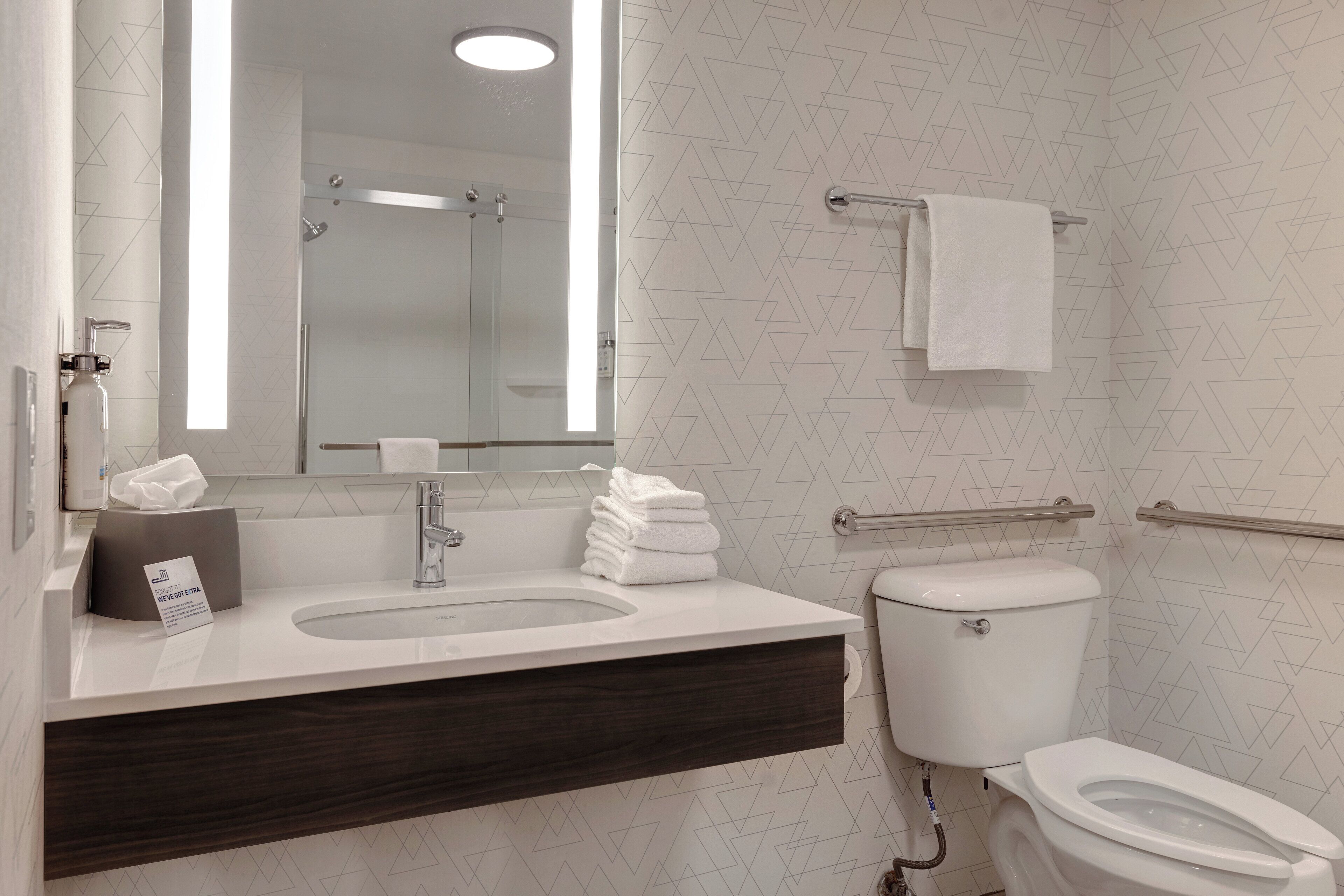 A modern bathroom with a white sink, mirror, and toilet. There is a towel rack above the toilet and a towel on it. A tissue box is on the sink counter.