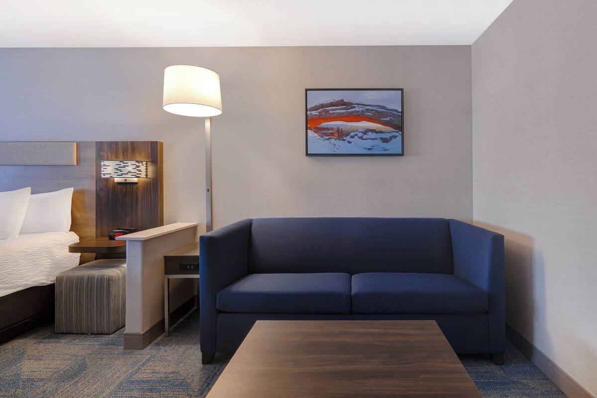 Una habitación de hotel moderna con cama, un sofá azul, una mesa de centro de madera, una lámpara y un cuadro en la pared.
