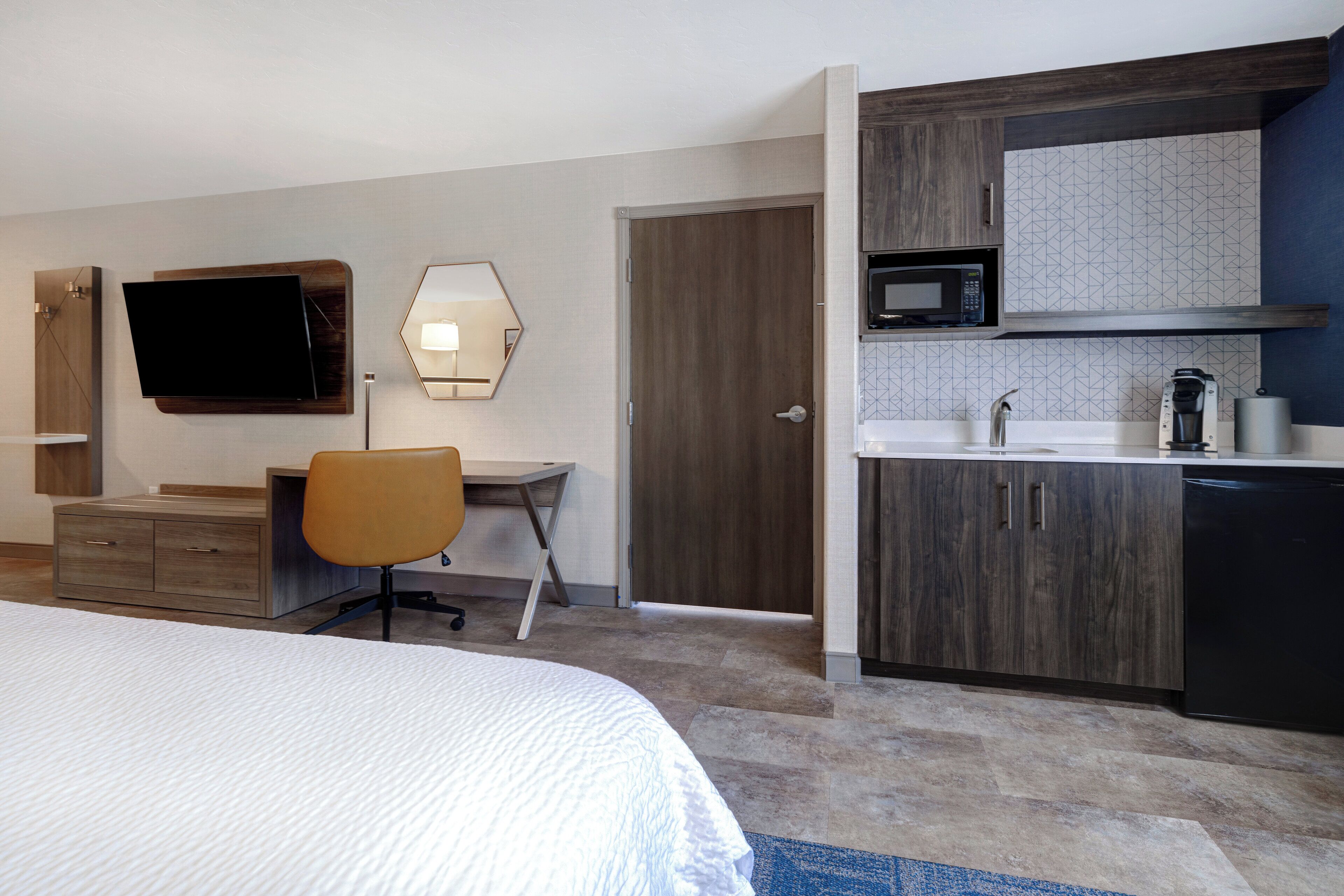 A hotel room with a bed, a desk, a chair, a television, a microwave, and a kitchenette.