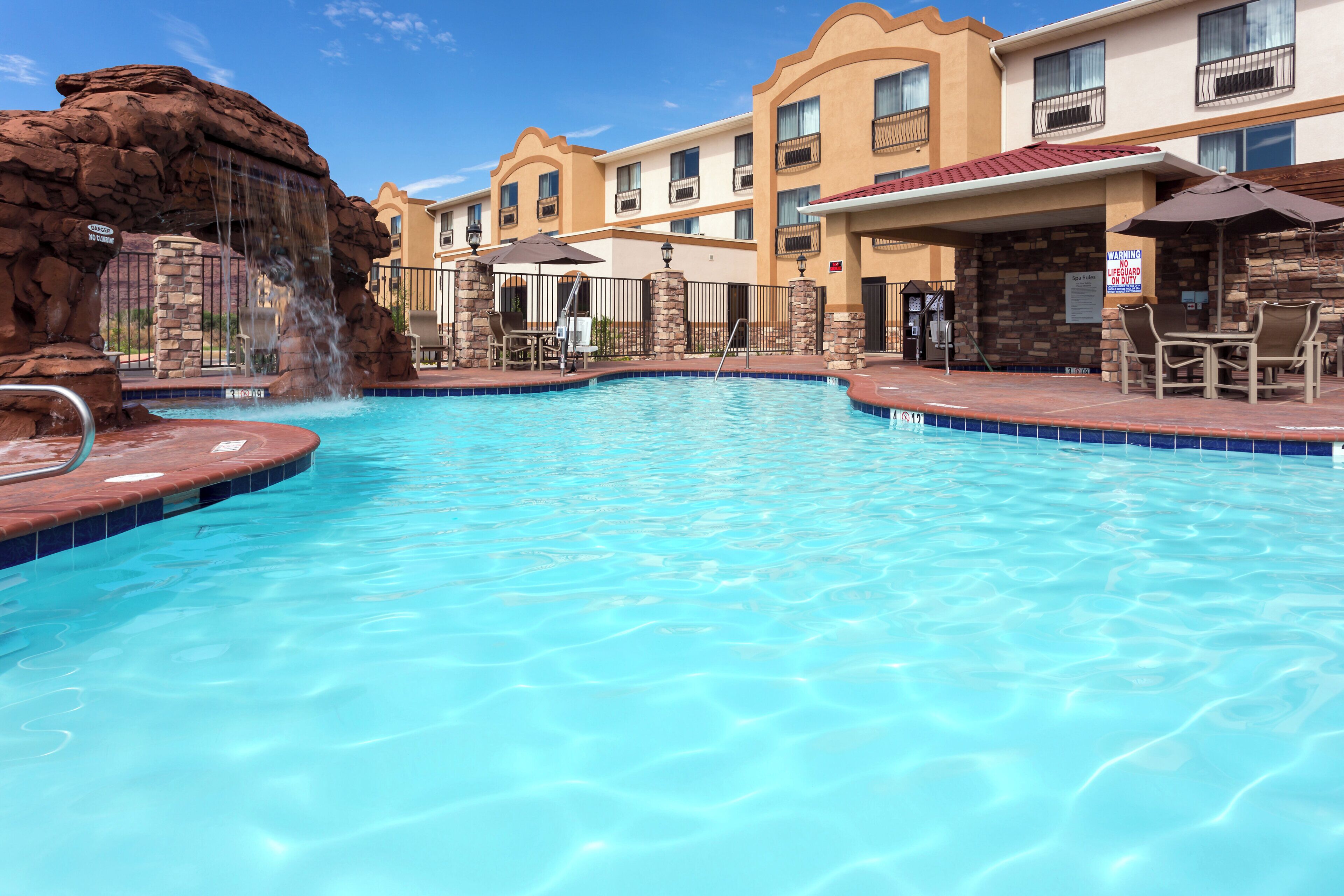 A swimming pool with a waterfall feature, surrounded by lounge chairs and a building with multiple balconies.