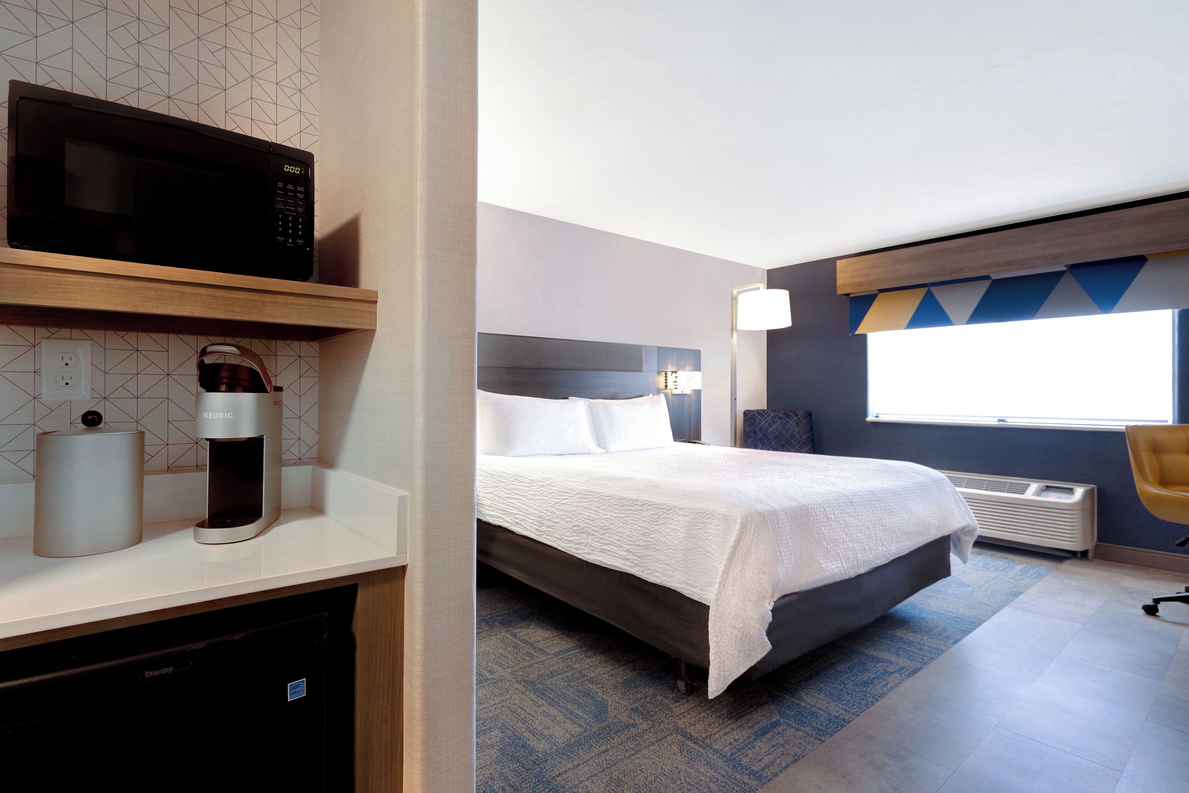 A hotel room with a bed, a microwave, a coffee maker, and a desk with a lamp.