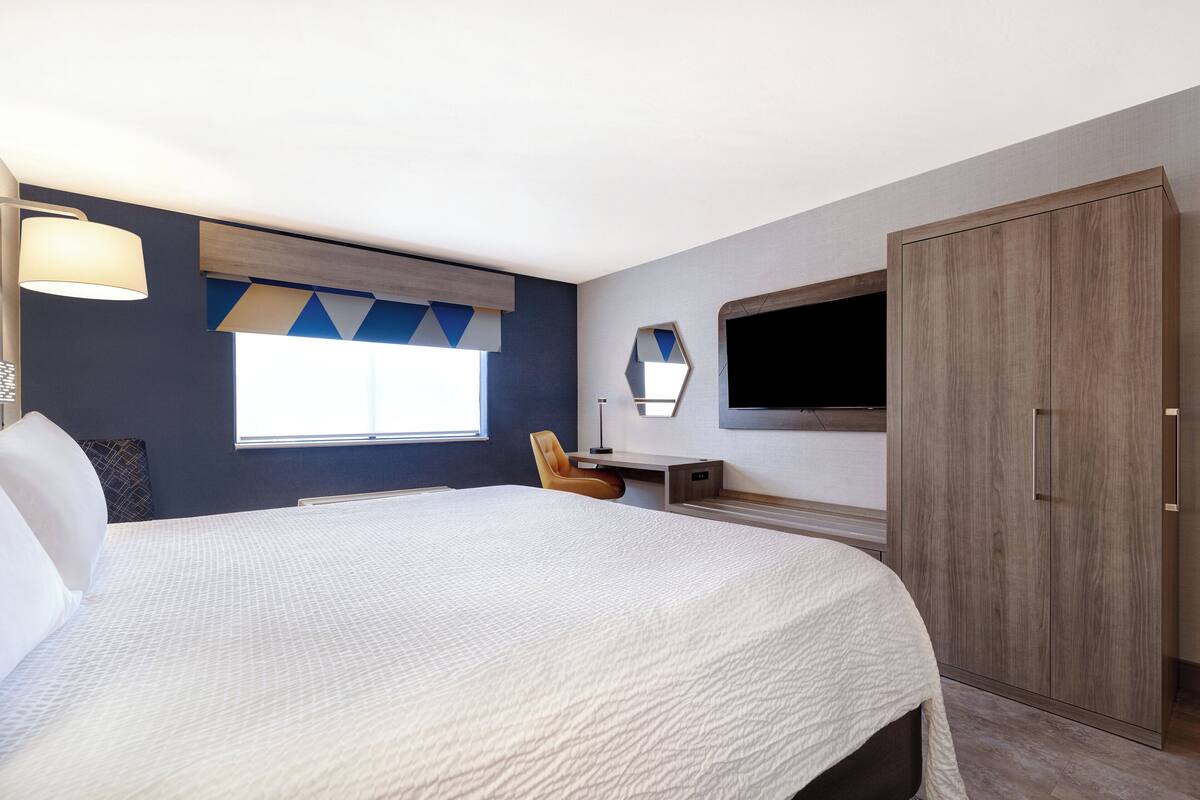 Habitación de hotel con una cama grande, un escritorio, una silla, un armario y un televisor montado en la pared.