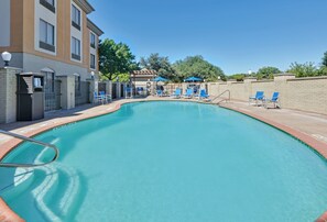 A swimming pool with blue chairs and umbrellas, surrounded by a brick wall and a building with multiple windows.