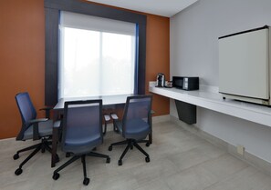 Uma sala de conferência com uma grande mesa, quatro cadeiras de escritório, um quadro branco, uma máquina de café e uma televisão.