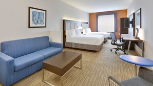 A hotel room with a blue sofa, a wooden coffee table, a bed with white linens, a desk with a chair, and a large window. - Holiday Inn Express Hotel & Suites Dallas - Duncanville by IHG (Duncanville)