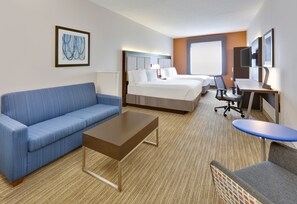 Quarto de hotel com sofá azul, mesa de centro de madeira, cama com roupa de cama branca, escrivaninha com cadeira e uma janela grande.