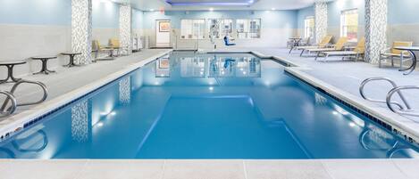 A large indoor swimming pool with multiple lanes, lounge chairs, and tables along the sides.