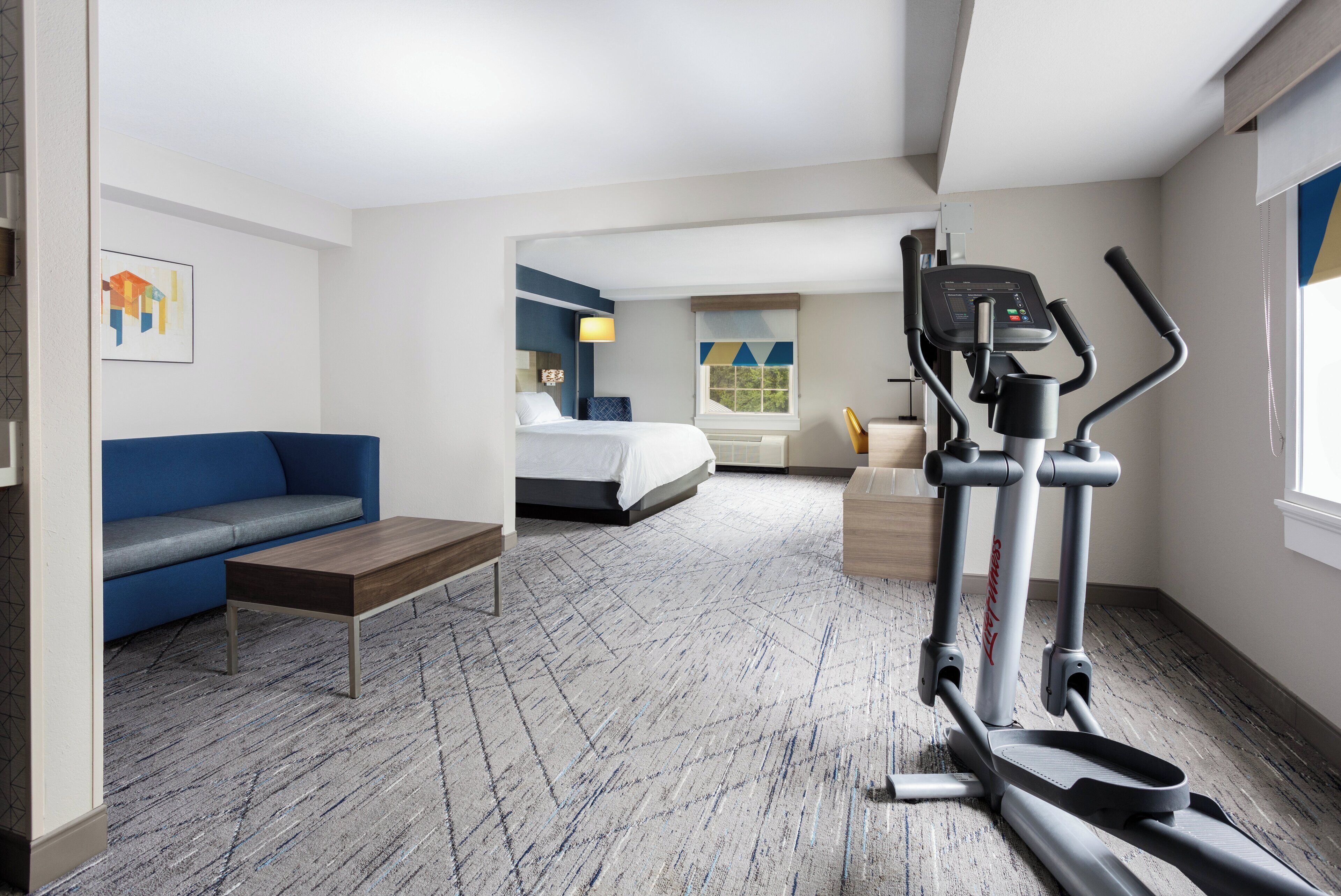 A hotel room with a bed, a blue sofa, a small table, a treadmill, and a window with a view.