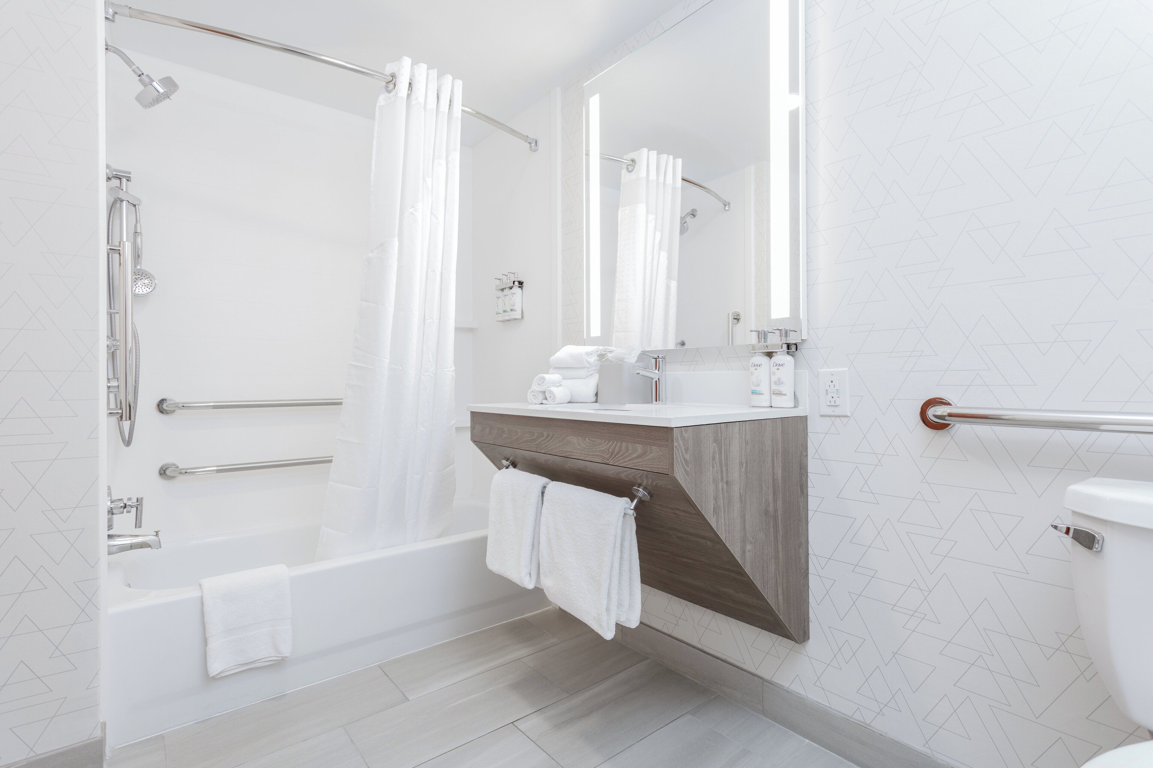 A modern bathroom with a bathtub, shower, and toilet. It features a wooden vanity, a towel rack, and a mirror.