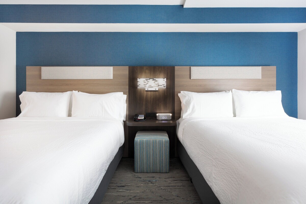 A hotel room with two beds, a nightstand with a clock, and a small ottoman.